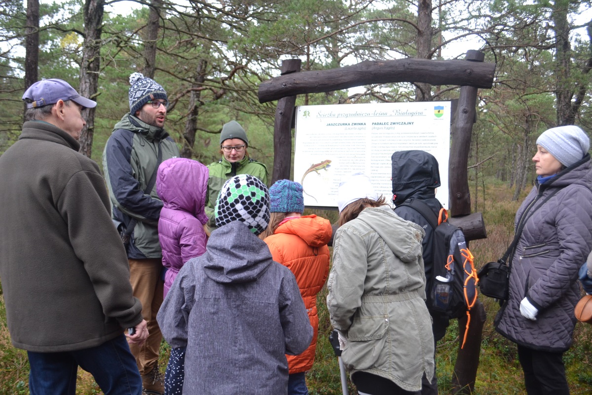 Warsztaty edukacyjne dla społeczności lokalnej w rezerwacie przyrody Białogóra; przy tablicy informacyjnej stoi grupa osób, słucha wykładowcy