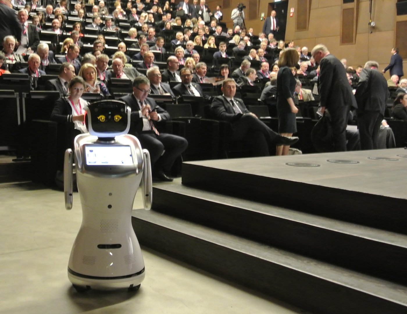 na pierwszym planie widoczny jest stojący robot humanoidalny. W tle siedzące osoby z publiczności.